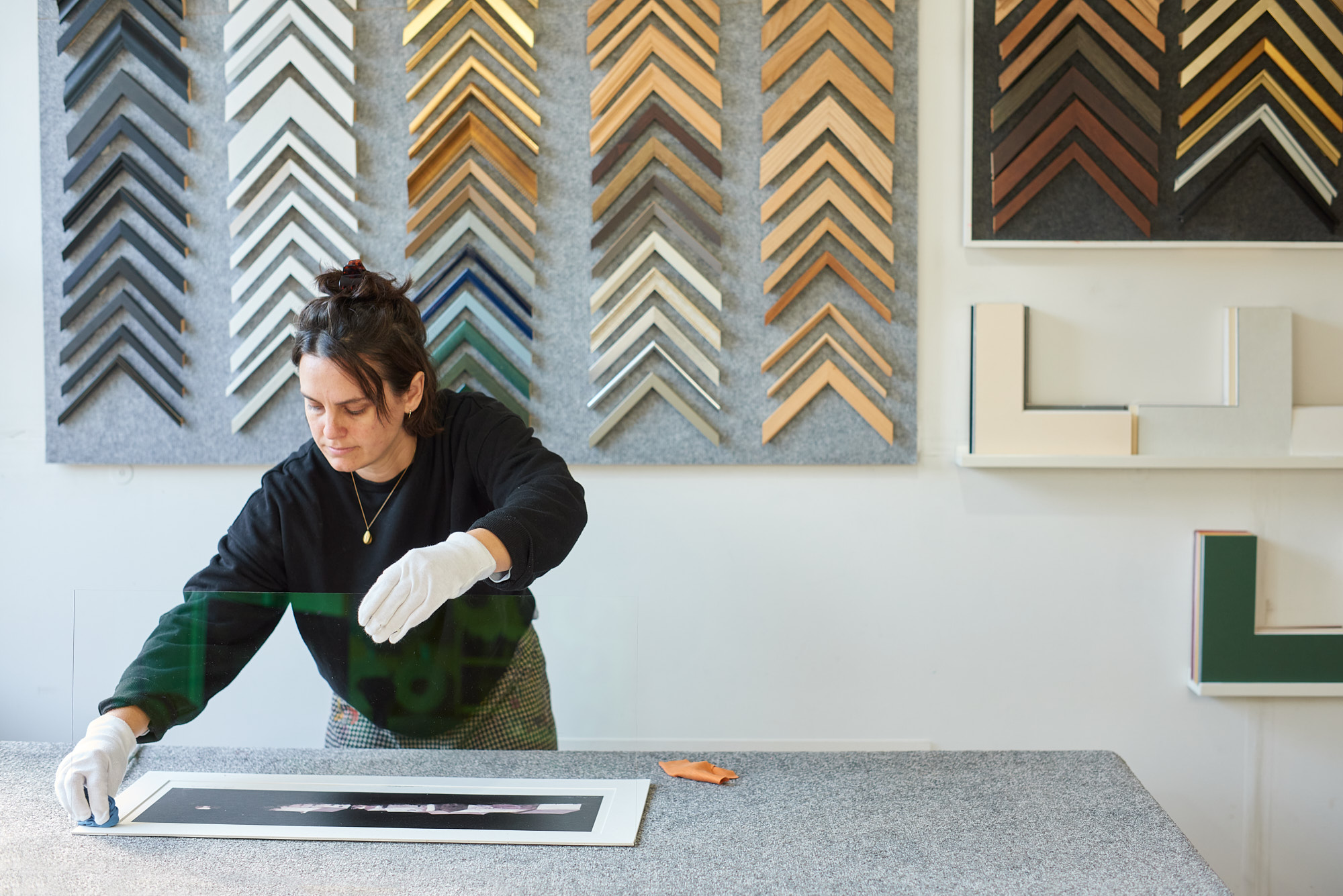 Photograph of framer wearing a black sweatshirt and white gloves supporting a piece glass and dusting mounted artwork with cloth